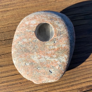 May include: A smooth, grey and pink stone with a round shape and a small, round hole in the centre. The hole is covered with a metal mesh.