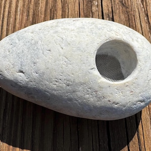 May include: A pale grey, oval-shaped stone with a large, circular opening. The opening reveals a mesh screen. The stone is resting on a wooden surface, and the lighting creates a shadow.