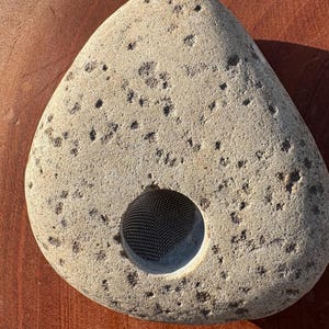 May include: A light grey, triangular stone with a large, circular hole. The stone has a rough, textured surface with small, dark spots. The hole appears to have a mesh lining. The stone is displayed on a wooden surface.