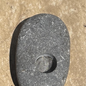 May include: A smooth, oval-shaped gray stone featuring a circular cutout in the center. The cutout is fitted with a fine, silver-colored metal mesh screen. The stone has a natural, speckled texture with variations in shade across its surface.