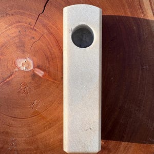 May include: A light beige rectangular object with a circular opening. The object has a textured surface and is set against a wood grain background. The object appears to be a pipe or a similar smoking accessory.