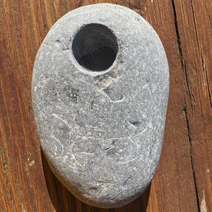 May include: A grey, oval-shaped stone with a circular opening. The stone has a textured surface and a dark mesh material visible within the opening. The stone is a natural, organic item.