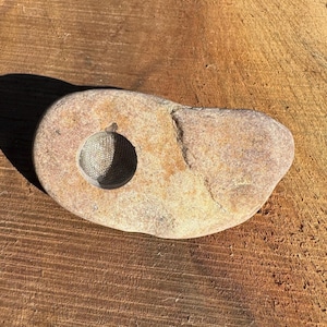 May include: A smooth, light brown stone with a round hole in the centre. The hole is lined with a silver mesh.