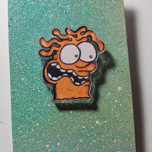 May include: A bright orange cartoon character with wild hair and big eyes is printed on a green background with white speckles. The character has a surprised expression and an open mouth. The background has the number 8/20 written in black ink.