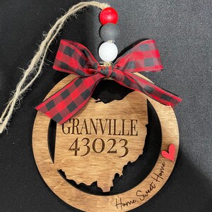 Op de afbeelding: Houten ornament in de vorm van de staat Ohio met de tekst "Granville 43023" en "Home Sweet Home" met een rood hart. De ornament is versierd met een rood-zwart geruite strik en rode, zwarte en witte kralen.