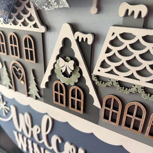 May include: A layered wooden winter-themed sign with a blue and gray color scheme. The sign features miniature houses, snowflakes, and the words "Welcome Winter" in white script. A blue bow with snowflake accents is at the top.