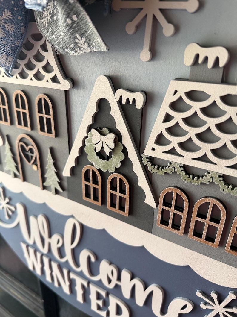 May include: A layered wooden winter-themed sign with a blue and gray color scheme. The sign features miniature houses, snowflakes, and the words "Welcome Winter" in white script. A blue bow with snowflake accents is at the top.