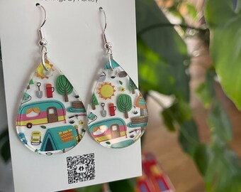May include: A pair of teardrop-shaped earrings with a white background and a colorful pattern of camping-themed illustrations, including a camper, trees, and a campfire. The earrings are made of clear acrylic and have silver hooks. The card they are displayed on reads "KMA DESIGNS Earrings by Ashley".