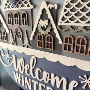 May include: A round, layered wooden sign with a winter village scene. The sign features houses with intricate cut-out details, a blue and white color scheme, and the words "Welcome Winter" in cursive. A decorative bow is at the top.