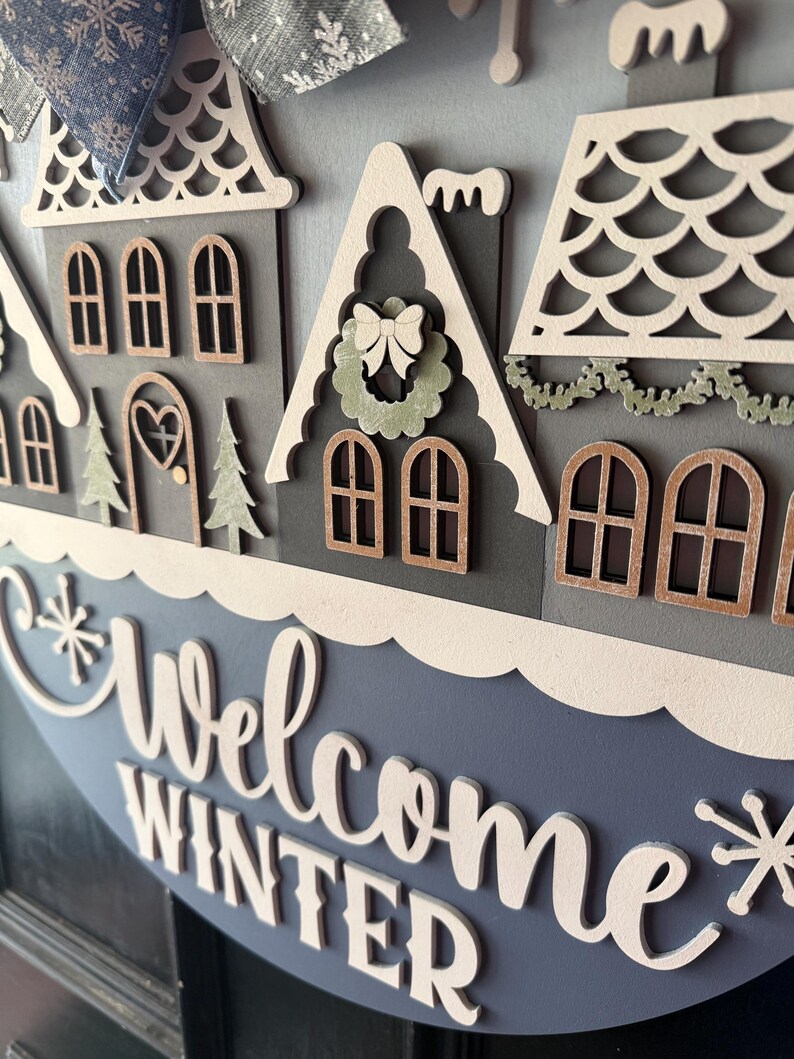 May include: A round, layered wooden sign with a winter village scene. The sign features houses with intricate cut-out details, a blue and white color scheme, and the words "Welcome Winter" in cursive. A decorative bow is at the top.