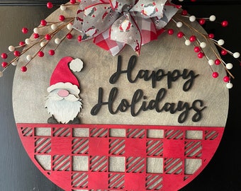 Puede incluir: Un colgador de puerta redondo de madera con un patrón de cuadros rojos y blancos en la mitad inferior. La mitad superior presenta un gnomo gris y blanco con un gorro rojo y las palabras "Happy Holidays" en letras negras. Un lazo de cuadros rojos y blancos con un estampado de gnomo gris y blanco está sujeto a la parte superior.