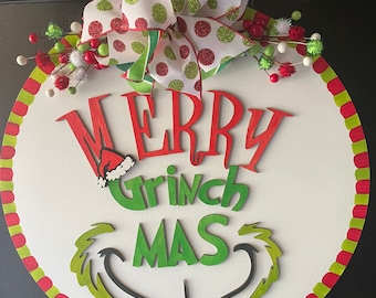 Może przedstawiać: Okrągły biały wieszak na drzwi z czerwonymi i zielonymi paskami wokół krawędzi. Słowa "Merry Grinchmas" są napisane zielonymi i czerwonymi literami z wzorem przypominającym Grincha poniżej. Czerwona i biała kokardka w kropki z zielonymi akcentami jest przymocowana do góry.