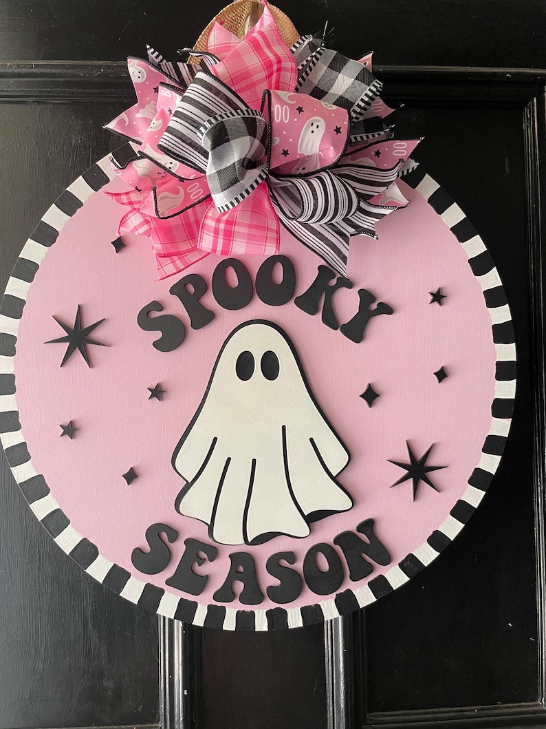 Puede incluir: Colgador de puerta de Halloween rosa y negro con un fantasma blanco y el texto "Spooky Season".