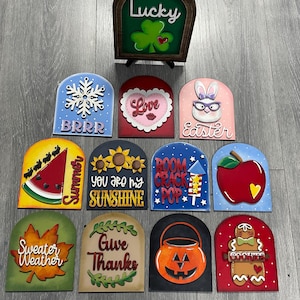 Könnte beinhalten: Eine Sammlung von saisonalen Holzschildern mit verschiedenen Designs und Texten. Enthält ein "Lucky"-Schild mit einem Kleeblatt, einen "BRRR"-Schneeflocke, ein "Love"-Herz, einen "Easter"-Hasen, eine Wassermelone, Sonnenblumen, ein Feuerwerk, einen Apfel, ein Blatt, einen Kürbis und einen Lebkuchenmann.