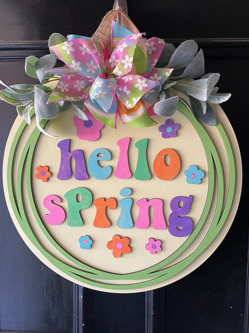 Pu&ograve; includere: Un appendiporta rotondo in legno con una ghirlanda verde e le parole "hello spring" in lettere colorate. La ghirlanda &egrave; decorata con un fiocco rosa e verde e piccoli fiori.
