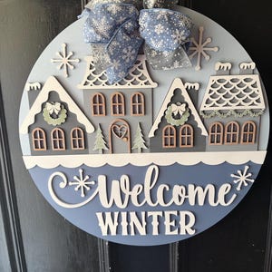 May include: A round wooden winter-themed door sign with a blue and white color scheme. The sign features a winter village scene with houses, snowflakes, and the words "Welcome Winter". A blue and gray snowflake bow is attached at the top.