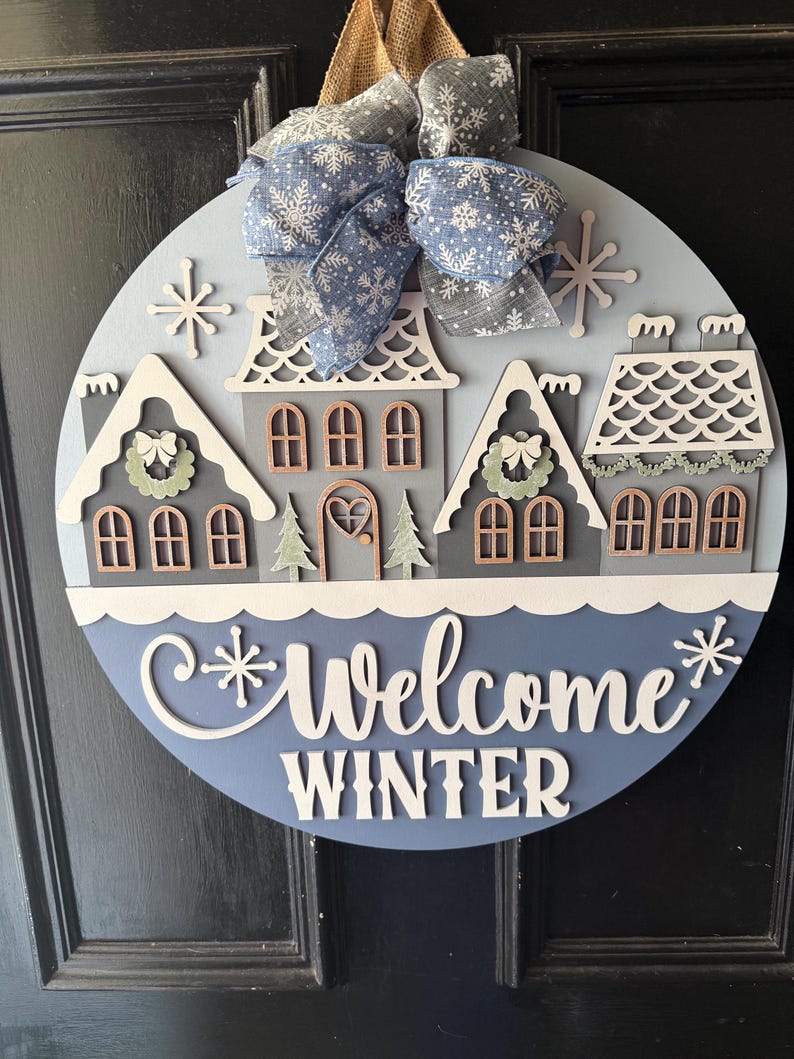May include: A round wooden winter-themed door sign with a blue and white color scheme. The sign features a winter village scene with houses, snowflakes, and the words "Welcome Winter". A blue and gray snowflake bow is attached at the top.