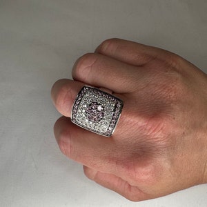 May include: A silver ring with a square-shaped setting, featuring a large pink gemstone surrounded by smaller white gemstones.