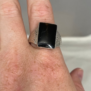 May include: A silver ring with a black rectangular stone set in the center. The band of the ring has a textured design.