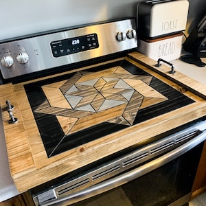 May include: A wooden stovetop cover with a geometric pattern in shades of brown, black, and gray. The cover is made of wood and has a natural finish. The cover is placed on top of a stainless steel stovetop.