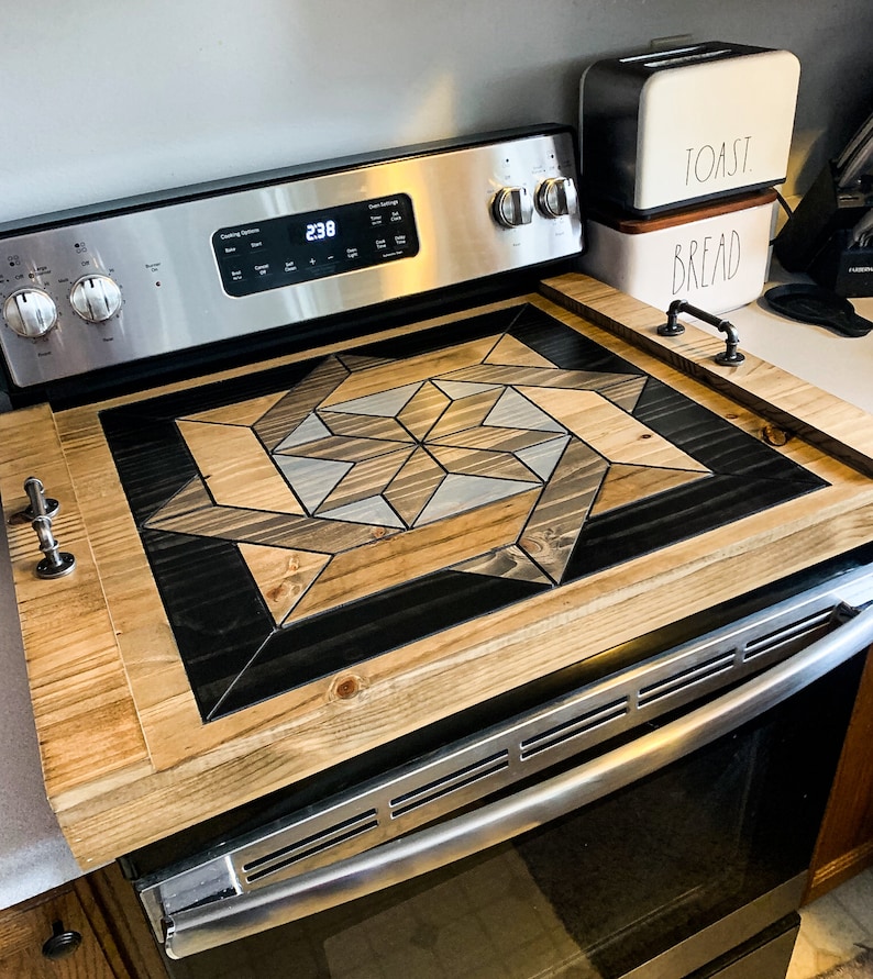 May include: A wooden stovetop cover with a geometric pattern in shades of brown, black, and gray. The cover is made of wood and has a natural finish. The cover is placed on top of a stainless steel stovetop.
