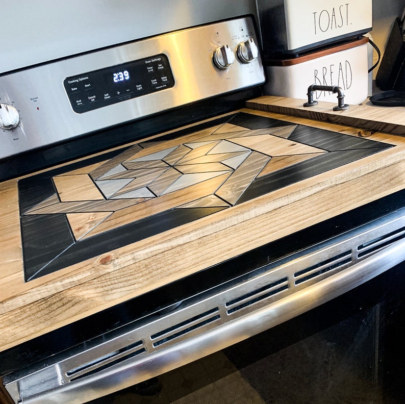 May include: A close-up of a wood stovetop cover with a geometric pattern in shades of brown, black, and white. The cover is placed over a stainless steel oven with a black trim.