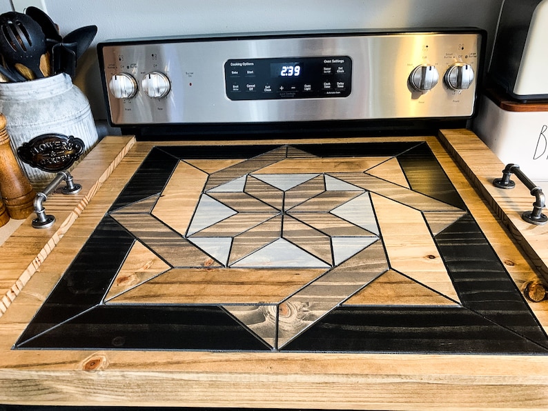 May include: A wooden stovetop cover with a geometric design in shades of brown, black, and gray. The cover is made of individual pieces of wood that are arranged in a starburst pattern. The cover is sitting on top of a stainless steel stovetop.