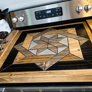 May include: A wooden stovetop cover with a geometric design in shades of brown, black, and gray. The cover has two metal handles on the sides.
