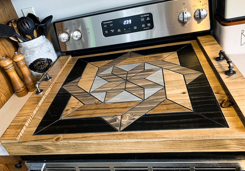 May include: A wooden stovetop cover with a geometric design in shades of brown, black, and gray. The cover has two metal handles on the sides.