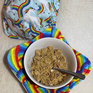 Puede incluir: Un tazón de cerámica blanco lleno de cereal se encuentra sobre un cojín de tazón de tela de arcoíris. El cojín tiene un patrón de arcoíris y unicornios.