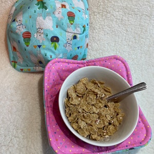 Puede incluir: Un salvamanteles azul claro con estampado de llama y un salvamanteles rosa con lunares que sostiene un cuenco blanco lleno de cereales. Una cuchara plateada descansa en el cuenco. El salvamanteles está acolchado.