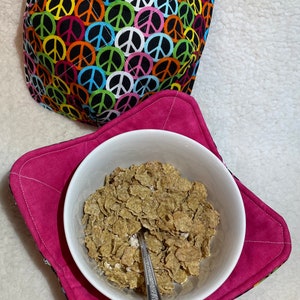 Puede incluir: Un calientaplatos rosa con un patrón de signo de paz en la parte inferior y un tazón blanco lleno de cereal y una cuchara dentro.