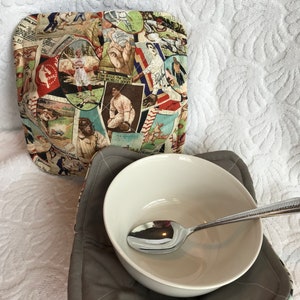 Puede incluir: Un calientaplatos acolchado gris con un estampado de temática de béisbol. Un tazón blanco con una cuchara de plata está dentro del calientaplatos.