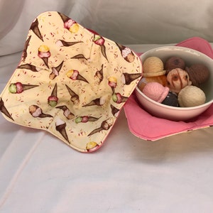 Puede incluir: Un calientaplatos de tela rosa y amarillo con un patrón de conos de helado. El calientaplatos sostiene un tazón blanco lleno de diferentes sabores de helado.