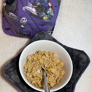 Puede incluir: Un tazón blanco lleno de cereal se encuentra sobre un cojín de tazón de tela negro y morado con una cuchara de plata en su interior. El cojín del tazón tiene un estampado de Pesadilla Antes de Navidad.