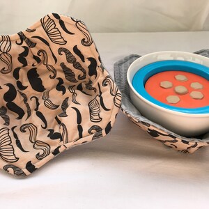 Puede incluir: Dos calientaplatos de tela beige con un patrón de bigote negro y blanco. Un calientaplatos está doblado y el otro sostiene un tazón blanco con un anillo interior azul y naranja.