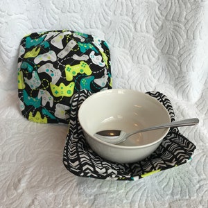 Puede incluir: Dos calientaplatos negros y blancos. Un calientaplatos tiene un fondo negro con un patrón de mandos de videojuegos verdes y blancos. El otro calientaplatos tiene un patrón de chevron negro y blanco. Un cuenco blanco con una cuchara de plata dentro está sentado en el calientaplatos de chevron.