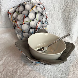 May include: A white ceramic bowl with a silver spoon inside, sitting on a gray and white fabric bowl cozy with a golf ball and tee design. The cozy is on a white textured surface.