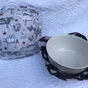 Puede incluir: Dos calientaplatos de tela con estampado de animales de granja. Un calientaplatos es blanco con estrellas rojas y animales de granja en blanco y negro. El otro calientaplatos es negro con animales de granja blancos. Ambos calientaplatos están hechos de tela de algodón.