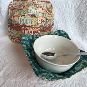 Puede incluir: Un juego de dos calientaplatos de tela azul verdoso y crema con un diseño vintage de la Ruta 66. Los calientaplatos presentan un mosaico de placas de matrícula con varios estados y números. Los calientaplatos son perfectos para mantener los cuencos calientes o proteger las superficies de los cuencos calientes.