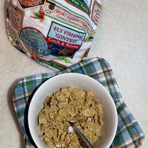 Puede incluir: Un tazón blanco lleno de cereal se encuentra sobre un mantel a cuadros. El tazón está al lado de una cuchara. En el fondo, hay una bolsa de tela con temática de pesca. La bolsa tiene las palabras "Fly Fishing Contest Catch & Release" impresas.