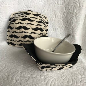 Puede incluir: Un tazón de cerámica blanco con un cojín de tazón de tela con estampado blanco y negro con un diseño de bigote repetido. El cojín del tazón está doblado hacia abajo para mostrar el forro de tela negro.