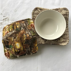 Puede incluir: Dos calientaplatos de tela acolchados con un patrón de camuflaje de bosque con un tazón blanco encima de uno de los calientaplatos.