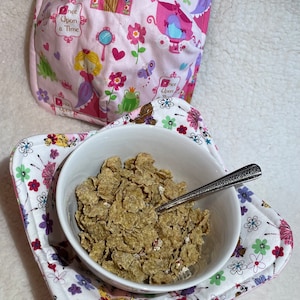 Puede incluir: Un tazón blanco lleno de cereal se encuentra sobre un mantel individual de tela rosa y blanco con un patrón floral. El mantel individual tiene las palabras "Once Upon a Time" impresas. Un calienta-tazón de tela rosa y blanco con un diseño de castillo y princesa está en el fondo.