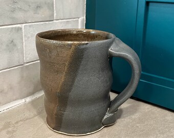 Puede incluir: Una taza de cerámica hecha a mano con un esmalte marrón y gris. La taza tiene un cuerpo redondeado y un asa.