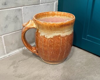 Puede incluir: Una taza de cerámica marrón con un esmalte marrón claro y un borde blanco. La taza tiene un asa y está sentada sobre una superficie gris.