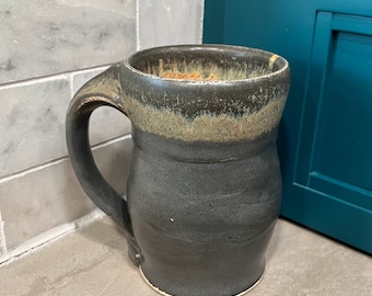Puede incluir: Una taza de cerámica negra con un esmalte marrón y beige. La taza tiene una base ancha y un cuerpo redondeado. Tiene una asa grande.