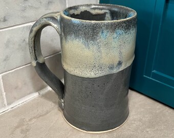 Puede incluir: Una taza de cerámica grande de color gris con un esmalte gris claro y azul. La taza tiene una asa ancha y redondeada.