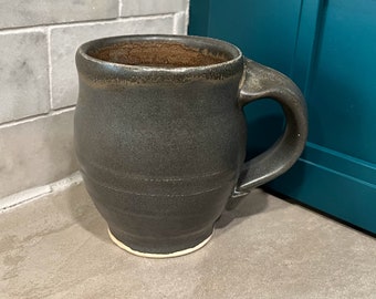 Puede incluir: Una taza de cerámica gris oscuro con un cuerpo redondeado y un borde ligeramente acampanado. La taza tiene un esmalte blanco en la parte inferior y un esmalte marrón en el interior.