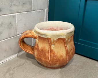Puede incluir: Una taza de cerámica marrón y beige con un glaseado blanco y un asa. La taza tiene un diseño texturizado y acanalado.